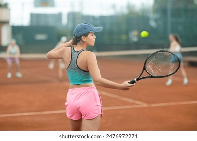 Emotional fit young girl, seen from behind, enthusiastically playing tennis outdoors, performing forehand to return ball to opponent court during doubles match with female partner.. - Powered by Shutterstock