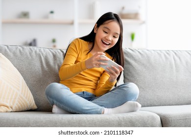 Emotional Fmale Kid Cute Long-haired Asian Girl Sitting On Sofa, Having Fun At Home, Using Smartphone And Laughing, Playing Video Game On Mobile Phone, Copy Space. Kids And Gadgets Concept