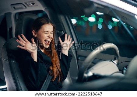 an emotional, frightened woman is sitting behind the wheel of a car in a black shirt, wearing a seat belt, expressing her emotions, spreading her arms to the sides and closing her eyes from fright. 