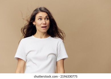 An Emotional, Funny Woman With Long, Well-groomed Hair Blowing In The Wind Is Standing In A White T-shirt With A Very Frightened Expression On Her Face. Horizontal Photo On A Beige Background