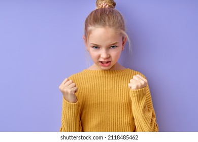 Emotional Girl In Yellow Shirt Over Isolated Purple Background Punching Fist To Fight, Aggressive And Angry Attack, Threat And Violence. Blonde Kid Girl Is Mad Raising Fist Frustrated And Furious