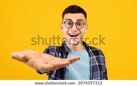 Emotional guy in casual outfit and glasses nerd outstretching big hand at camera, asking for something, posing over yellow studio background, panorama with copy space, creative image