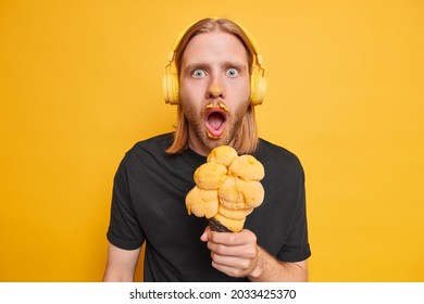 Emotional Guy With Red Long Hair Stares Shocked Finds Out Horible News Eats Sweet Summer Dessert Enjoys Favorite Songs From Playlist Isolated Over Yellow Background. People And Lifestyle Concept