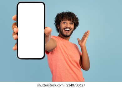 Emotional handsome curly young indian man in stylish pink t-shirt showing brand new cell phone with white empty screen and gesturing, sharing exciting online deal, blue background, mockup