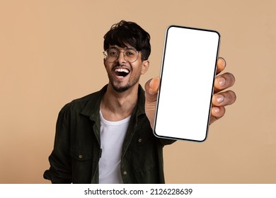 Emotional Handsome Hindu Guy Wearing Eyeglasses Showing Brand New Cell Phone With White Empty Screen And Smiling, Recommending Nice Online Offer, Mockup, Beige Studio Background