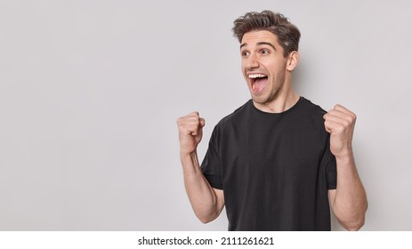 Emotional handsome man shakes clenched fists makes supportive gesture watches football match on TV dressed in casual black t shirt isolated over white background blank copy space. Supportive guy