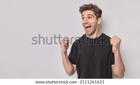 Emotional handsome man shakes clenched fists makes supportive gesture watches football match on TV dressed in casual black t shirt isolated over white background blank copy space. Supportive guy
