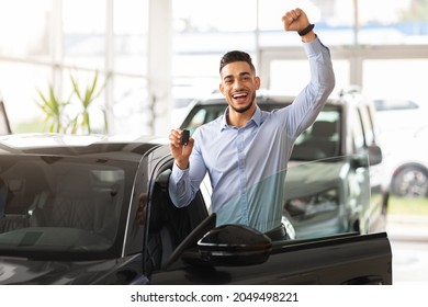 Emotional handsome middle-eastern guy raising hand up and smiling at camera, showing key from his brand new black car, copy space. Rich arabic man buying auto at luxury dealership salon