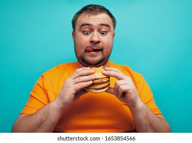 Emotional happy fat man on a blue background eating fast food hamburgers. Crazy food lover.