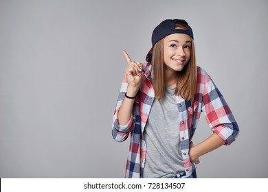 Emotional happy teen girl shrugging shoulders and pointing to the side up at empty copy space, over grey background.