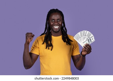 Emotional happy young african american guy in yellow showing bunch of cash dollars and gesturing, clenching fist, celebrating success over purple studio background. Gambling, trading, giveaway - Shutterstock ID 2249848003
