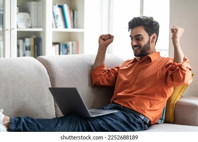 Emotional happy young middle eastern man in casual sitting on couch with legs up, raising hands up, celebrating success, guy trading on stock and markets while staying home, copy space, side view - Shutterstock ID 2252688189