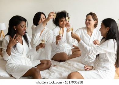 Emotional happy young mixed race girlfriends in white gowns toasting with white sparkling wine, celebrating birthday hen party at home or luxury spa hotel, listening to music, having fun together. - Powered by Shutterstock