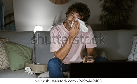 Emotional hispanic man crying while sitting on a couch indoors, wiping tears with a tissue.