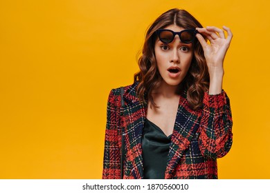 Emotional lady in striped outfit looking in surprise into camera on isolated backdrop. Curly girl with black sunglasses posing on yellow background..