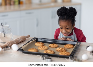 Emotional little black girl in apron and flour on her face looking at fresh homemade cookies on tray, cute african american child enjoying baking at home, happy with delicious result, copy space - Powered by Shutterstock