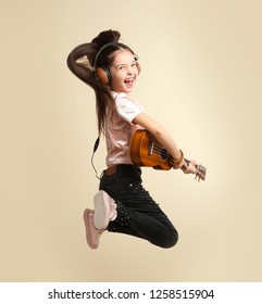 Emotional little girl with headphones playing guitar on color background