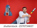 Emotional man hitting rocket shaped pinata with stick on red background