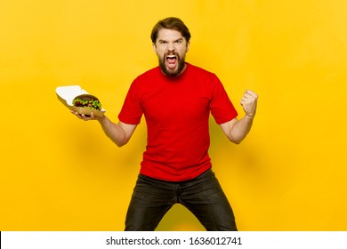 An emotional man in a red t-shirt and pants holds a hamburger in his hand on a yellow background - Shutterstock ID 1636012741