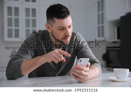 Emotional man with smartphone at table in kitchen. Online hate concept