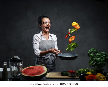 Emotional Mature Female Chef Tossing Chopped Vegetables From A Pan. Healthy Food Concept. Studio Photo On A Dark Background