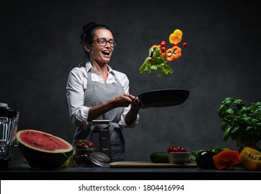 Emotional mature female chef tossing chopped vegetables from a pan. Healthy food concept. Studio photo on a dark background