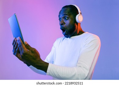 Emotional Middle Aged Black Man In Casual Outfit With Wireless Headset Looking At Digital Tablet Screen Over Neon Studio Background, Watching Cool Advertisement Online, Using Nice Entertaining App