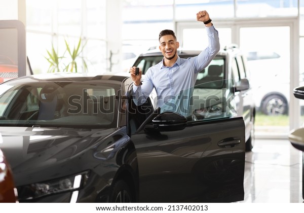 Emotional middle\
eastern guy buying car of his dream, happy male customer standing\
by nice shiny black auto at automobile dealership, showing key and\
raising hand up, copy\
space