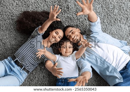 Emotional middle-eastern family father, mother and little daughter bonding on floor, cheerfully smiling and raising hands up, top view, closeup. Family, parenthood, parents and kids concept