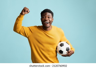 Emotional Millennial Black Guy In Casual With Soccer Ball Posing On Blue Studio Background, African American Young Man Football Fan Going To Game, Raising Fist Up, Copy Space. Sport Concept