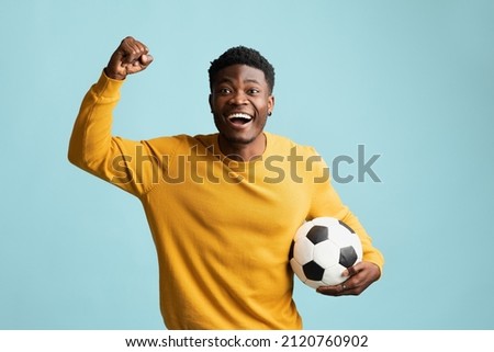 Emotional millennial black guy in casual with soccer ball posing on blue studio background, african american young man football fan going to game, raising fist up, copy space. Sport concept