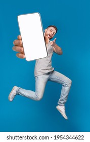 Emotional millennial redhaired guy pointing finger at camera, jumping with mobile phone on blue studio background, mockup for mobile app on white screen. Creative collage with website template