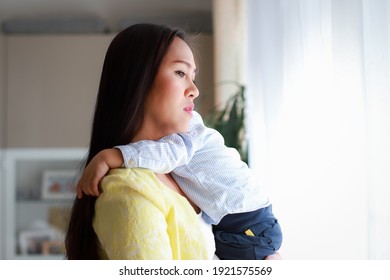 Emotional Mother Holding Baby In Her Arms Looking Outside The Window Feeling Tired Or Sad. Young Asian Woman Stressed With Her Kid. Housewife Overwork, Social Distancing Concept.