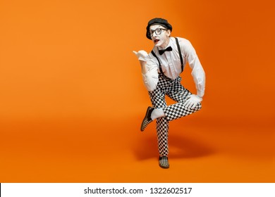 Emotional pantomime with white facial makeup showing empty space on the orange background, advertising something - Shutterstock ID 1322602517