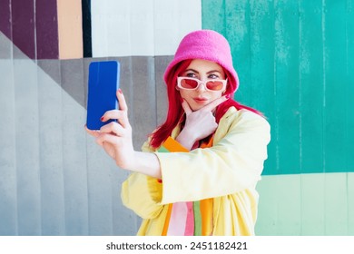Emotional pink hair woman taking selfie photo on graffiti background. Hipster fashion woman in bright clothes, pink sunglasses, bucket hat taking selfie photo on the phone camera. Influencer, blogger
