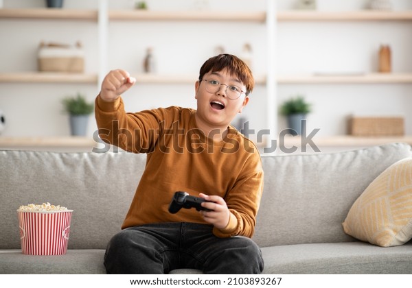 Emotional plump asian boy teenager sitting on couch\
in living room, holding joystick, playing handheld video game,\
eating popcorn, enjoying weekend at home, kids entertaining\
concept, copy space