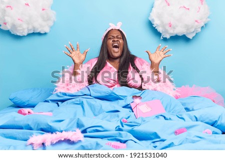 Emotional plump dark skinned Afro American woman raises palms shouts loudly dressed in dressing gown poses in bed with different items around isolated over blue background fluffy white clouds above