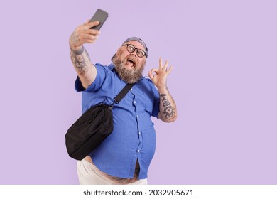Emotional plump hipster man with crossbody bag shows Ok gesture taking selfie with modern mobile phone on purple background in studio