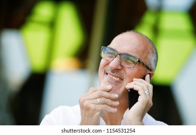 Emotional Portrait Of An Active And Sociable Mature European Man With Glasses And Bald Head Talking On The Smartphone With The Head And Rubbing His Chin With His Hand. Senior Hispanic Man. Lifestyle