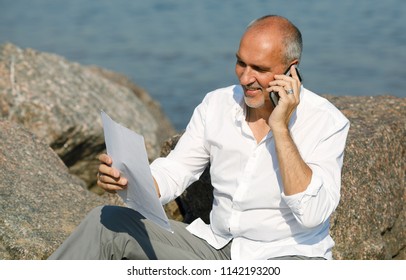 Emotional portrait of a business and positive mature European man reading a contract and discussing it on a smartphone sitting on a rocky beach during a summer holiday against the backdrop of the sea - Powered by Shutterstock