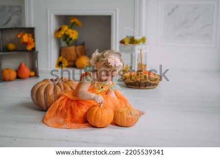 emotional portrait of a cute little happy 1 year old baby toddler kid girl in pumpkin costume is sitting on a pumpkin next to fireplace at home smiling. Halloween party concept. Easy costume idea. 