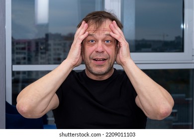 Emotional Portrait Of An Elderly Man Of 45-50 Years Old In A Black T-shirt, Holding His Hands To His Head, Neutral Background.