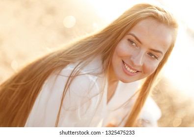 Emotional portrait of a happy and cheerful beautiful girl with blond hair looking with a smile at the beach lit by the sunset rays against the background of the sea.Summertime.Summer vacation