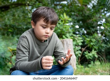 Emotional Portrait Of Lonely Kid Sitting Alone On Grass In The Garden, Sad Childplaying With Leaves And Looking Down Deep In Through, A Boy Playing Alone In Green Nature Park, Mental Health