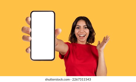 Emotional pretty young arab woman in red t-shirt showing brand new cell phone with white empty screen and gesturing over yellow studio background, online offer, mockup, panorama - Shutterstock ID 2267311543