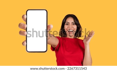 Emotional pretty young arab woman in red t-shirt showing brand new cell phone with white empty screen and gesturing over yellow studio background, online offer, mockup, panorama