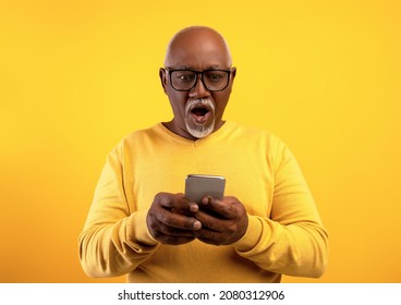 Emotional Senior African American Man In Glasses Looking At His Smartphone In Shock On Orange Studio Background. Surprised Elderly Black Male With Mobile Phone Amazed At Promo Sale Or Huge Discount