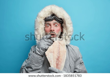 Emotional shocked frosty man dressed in winter outerwear holds frozen fish feels very cold during low temperature in northern place isolated on blue wall travels through blizzard. Male adventurer