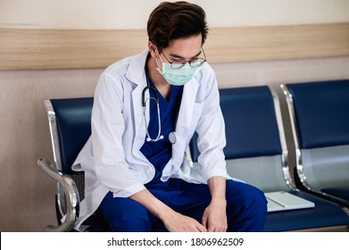 Emotional Stress Of Young Asian Man Doctor Being Exhausted And Worried About The Patient's Case While Sitting In The Hospital. Healthcare Worker Wearing Surgical Mask Looking Distraught And Tired. 