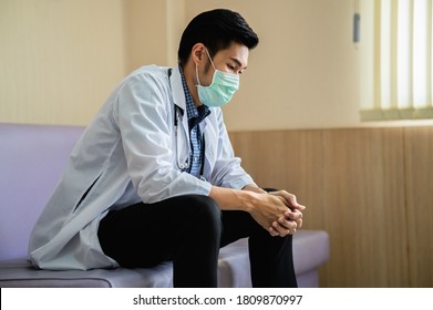 Emotional Stress Of Young Asian Man Doctor Being Exhausted And Worried About The Patient's Case While Sitting In The Hospital. Healthcare Worker Wearing Surgical Mask Looking Distraught And Tired. 